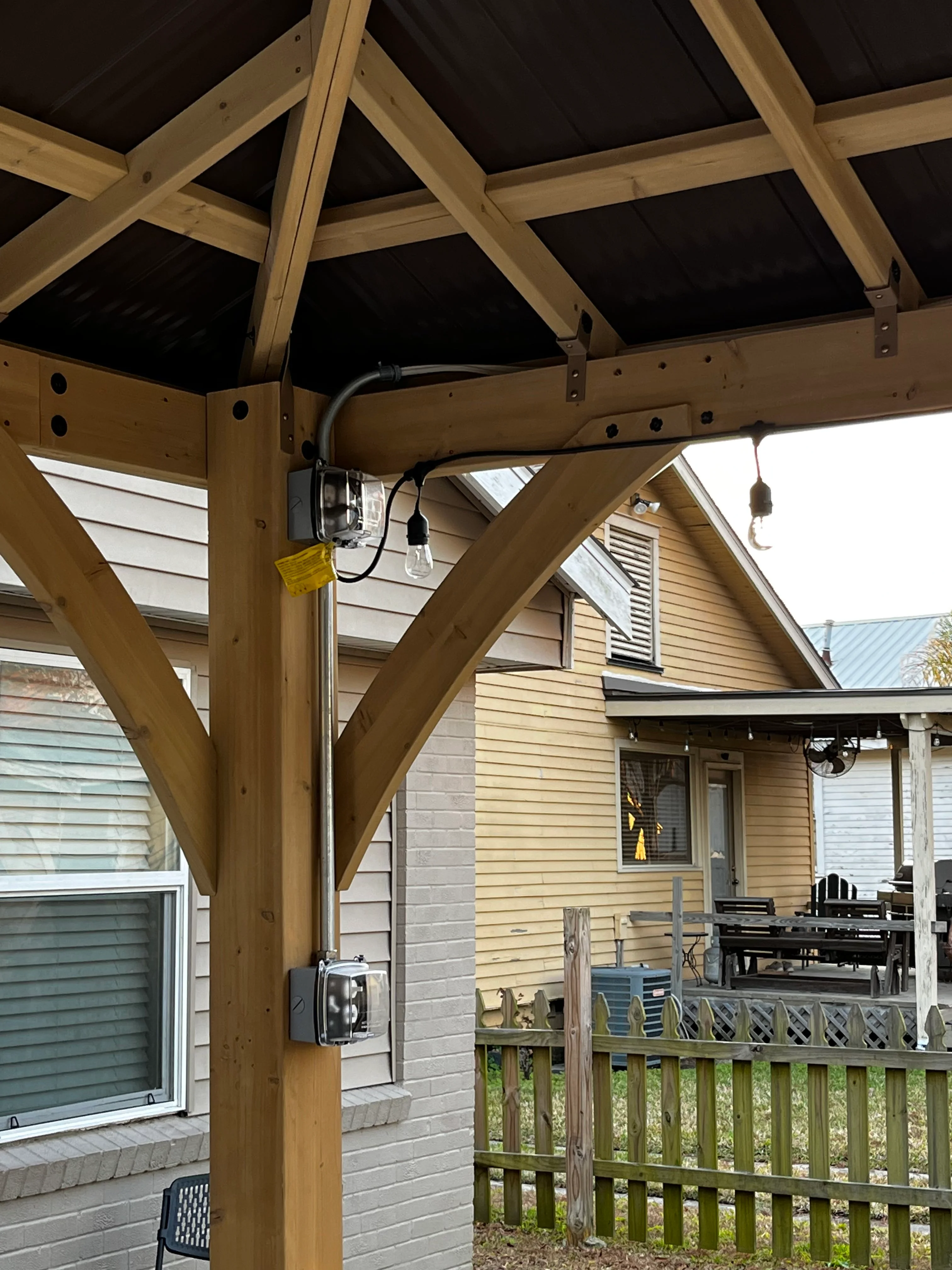 Timber pergola with exposed beams, black roof panels, and integrated electrical fixtures over residential patio