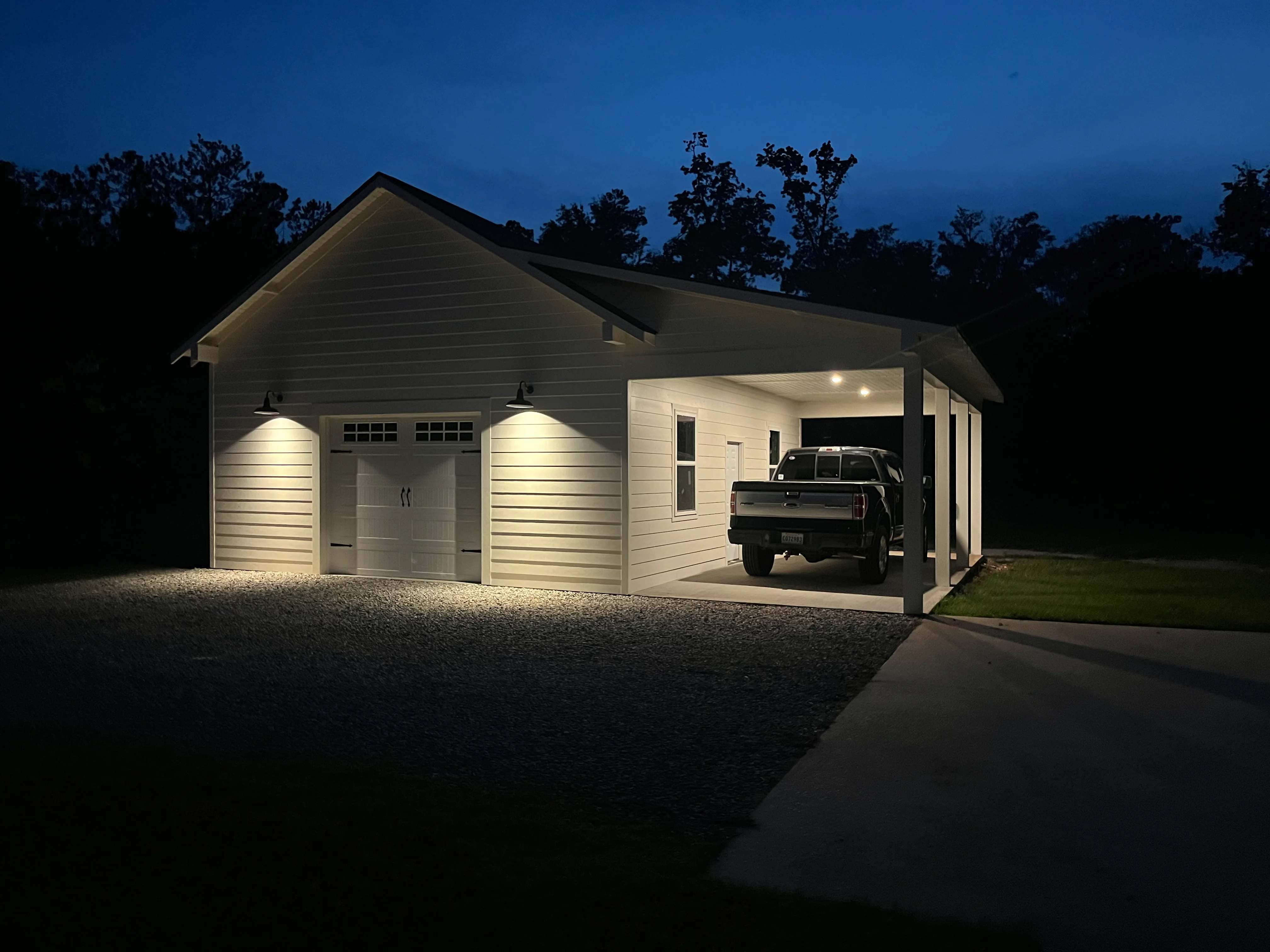 Illuminated two-car garage with carport at dusk, white siding, black roof, truck parked underneath