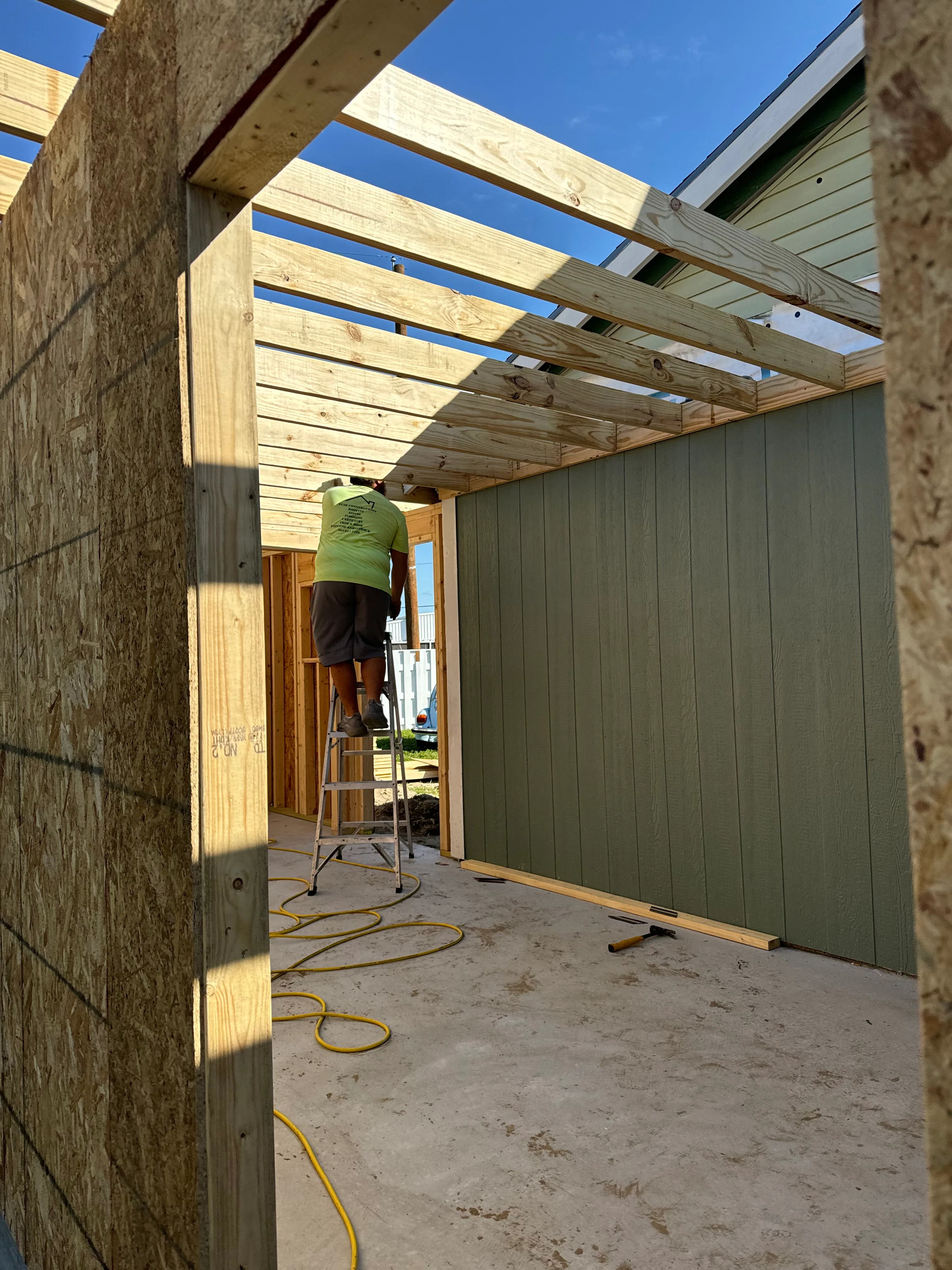 Worker installing wooden pergola beams over patio with brick walls and metal siding