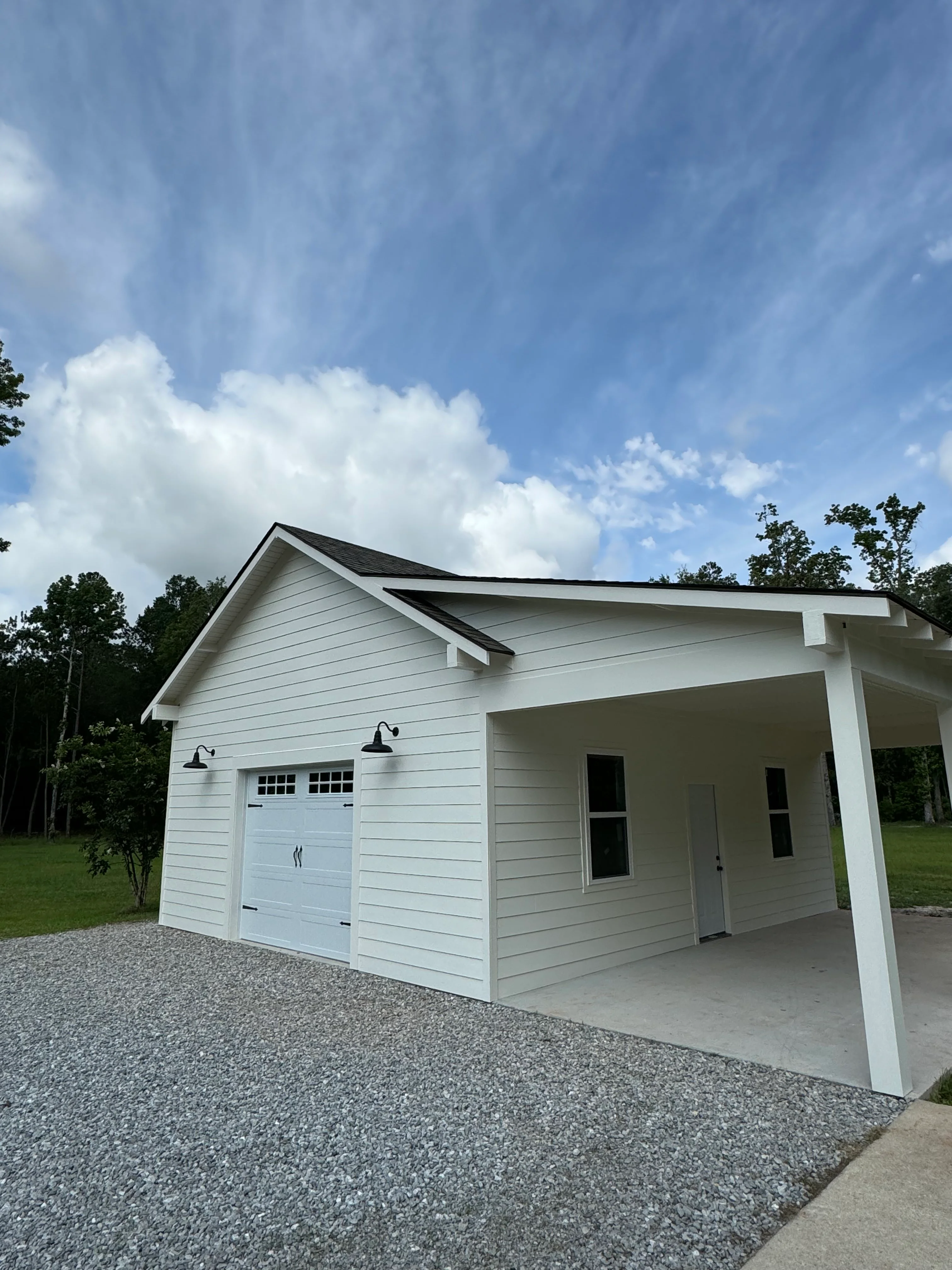 White garage with carport, black roof, and gravel driveway surrounded by trees