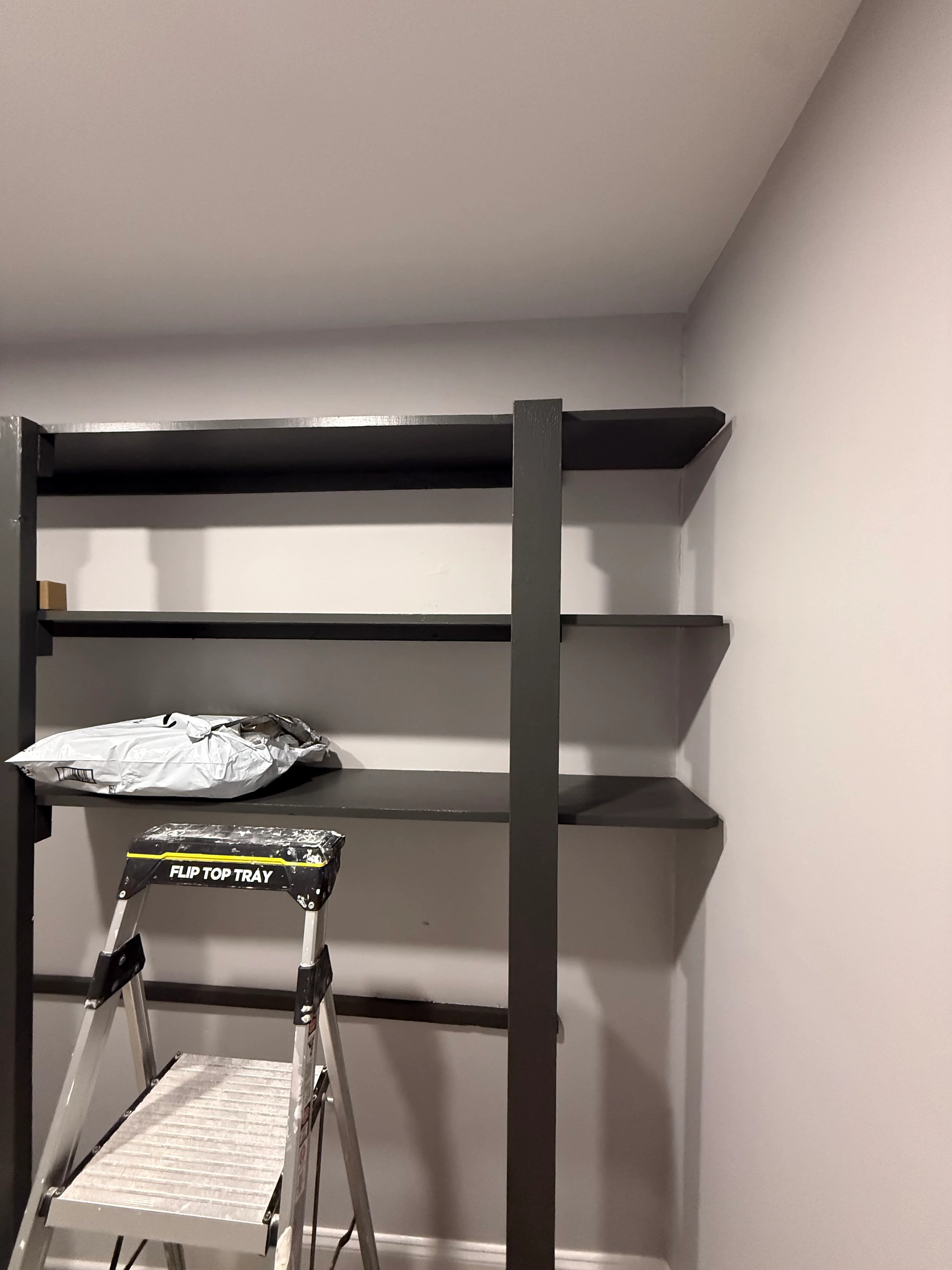 Black metal shelving unit with four shelves installed against white drywall in bedroom