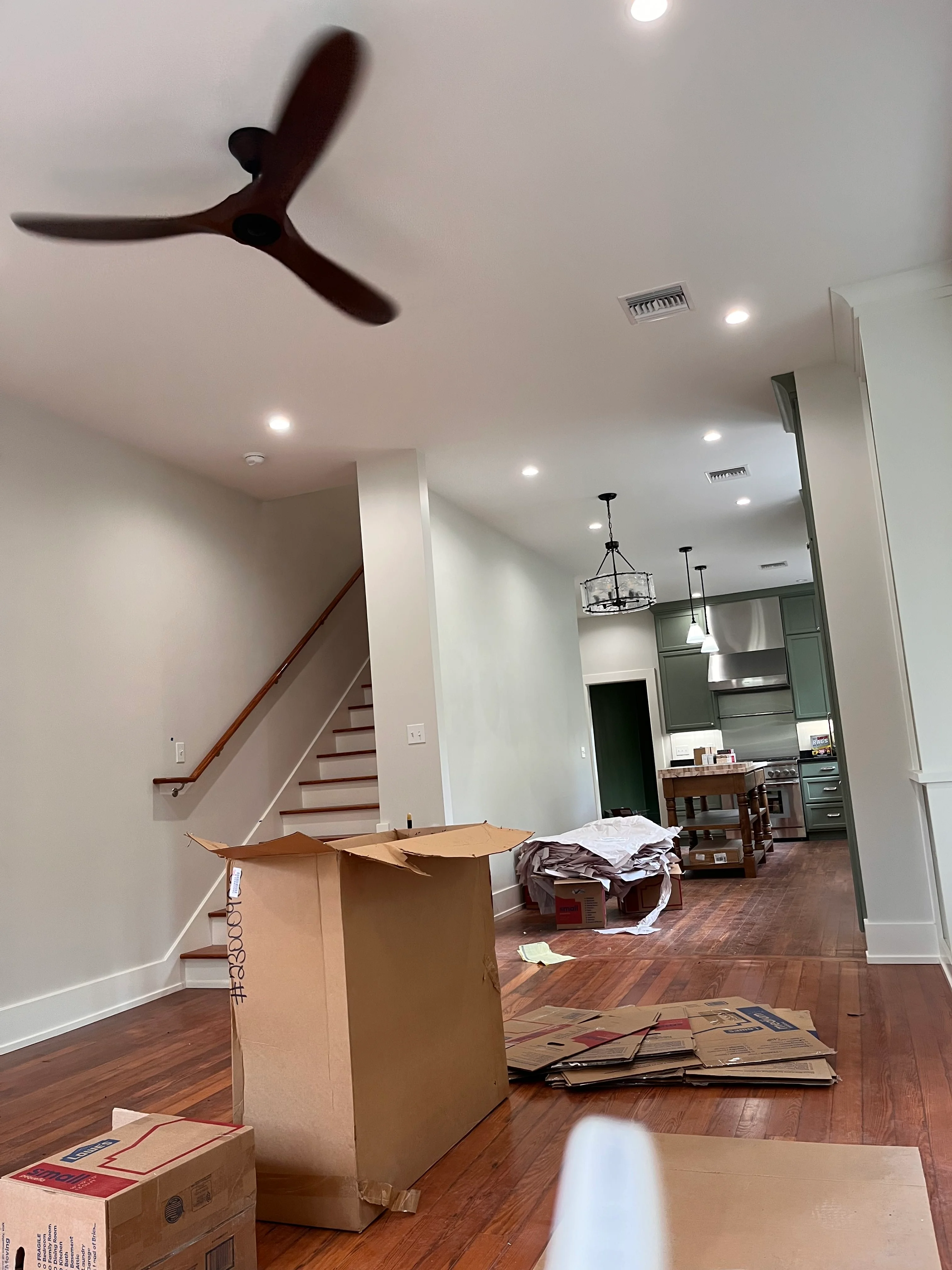 Interior renovation in progress showing hardwood floors, white walls, staircase, and kitchen with green cabinetry