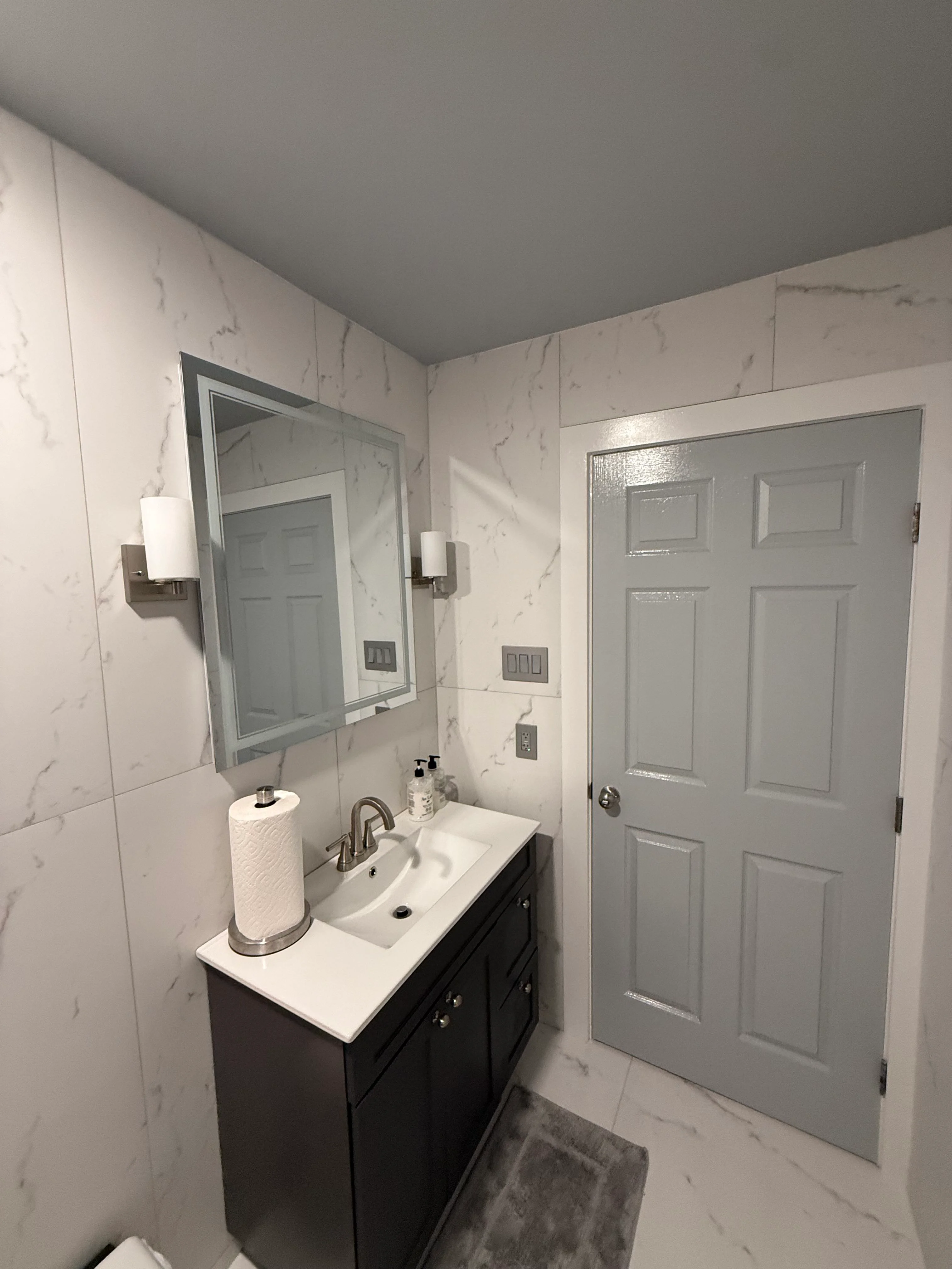 Modern bathroom with marble tile walls, dark vanity, and framed mirror with sconces