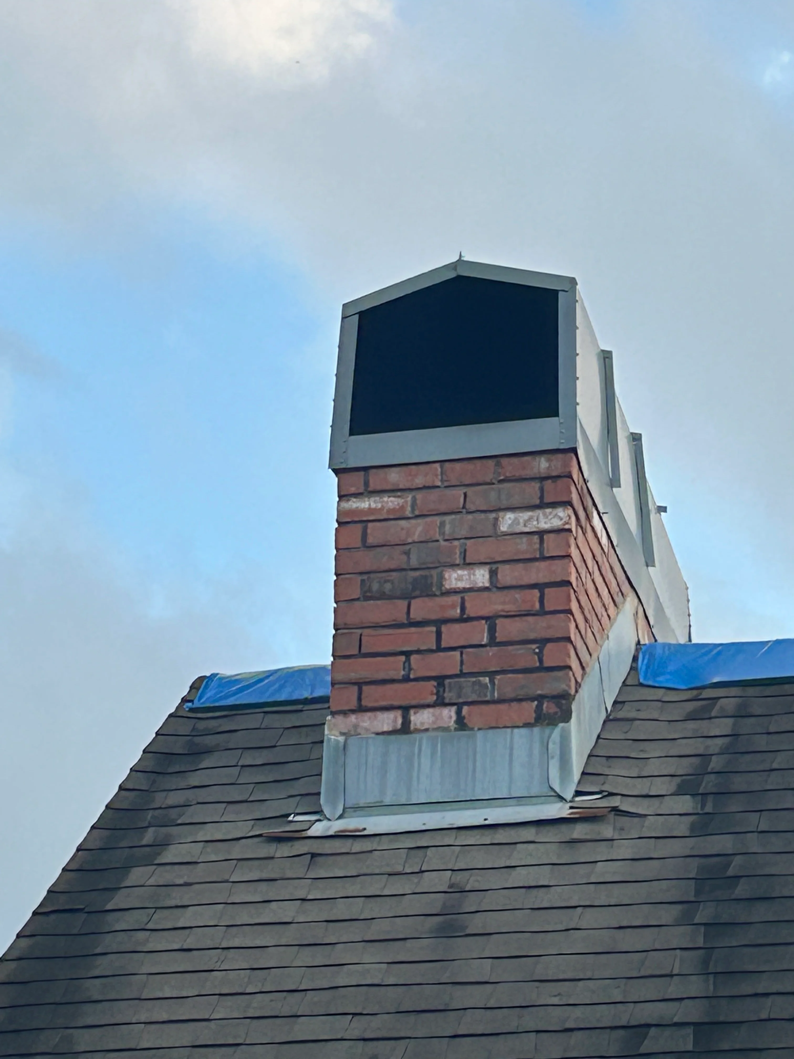 Newly rebuilt brick chimney with metal cap on slate roof with blue tarping